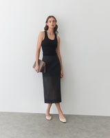 Woman in sleeveless top and skirt holding envelope clutch and standing against blank wall.