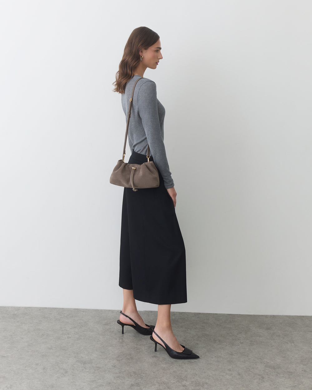 Woman standing sideways with shoulder bag and wearing a long skirt and heels.