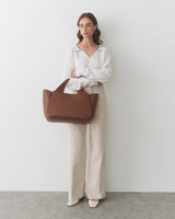 Woman holding a large handbag with both hands, wearing wide-leg pants and a long-sleeve top.
