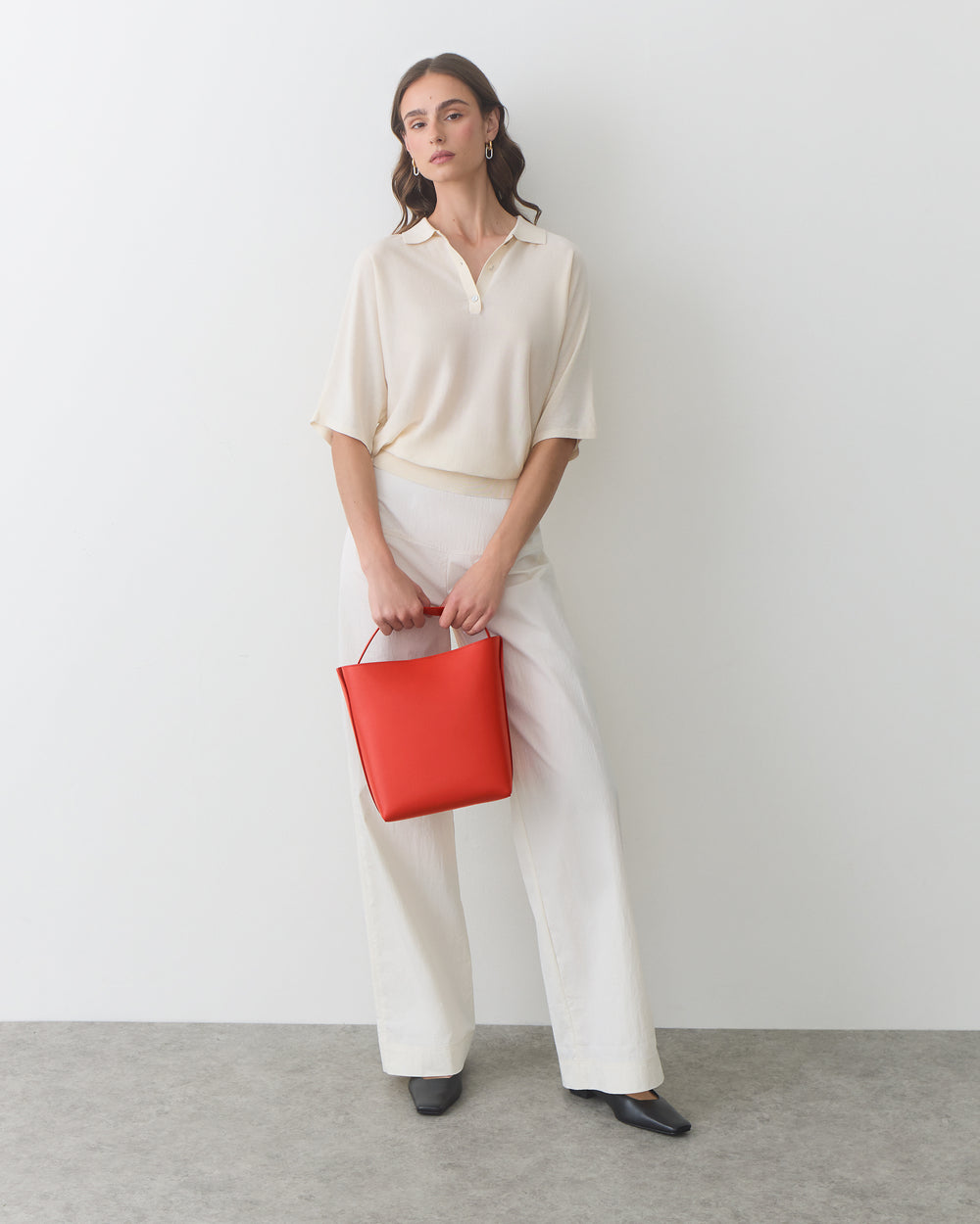 Person in relaxed outfit holding a handbag, standing against a plain background.
