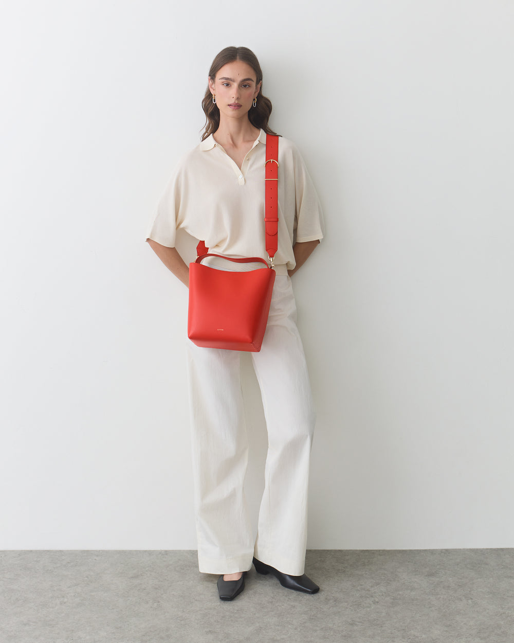 Person standing against a wall with a shoulder bag and hands in pockets.