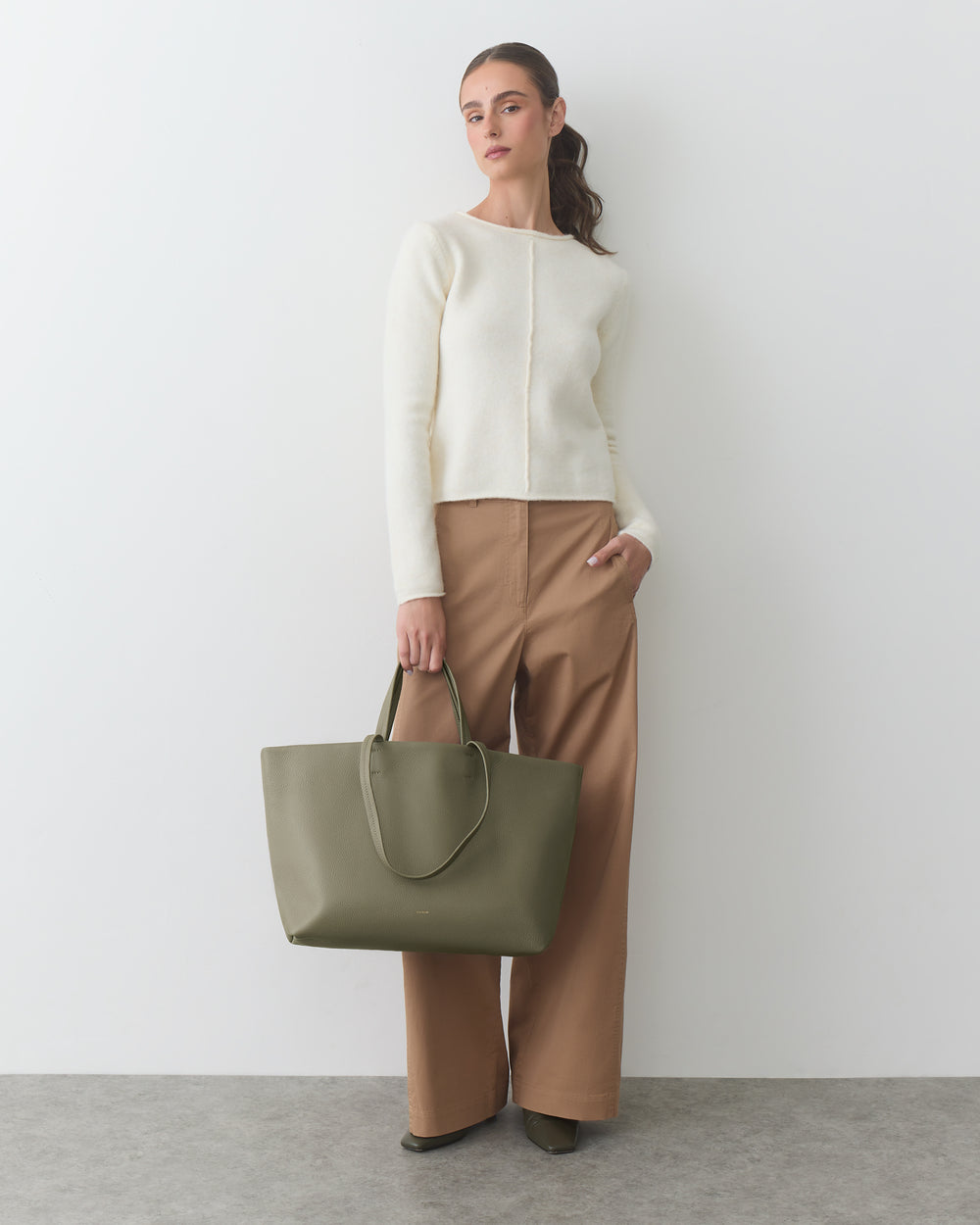 Woman standing with a large handbag, wearing a long-sleeve top and wide-leg pants.