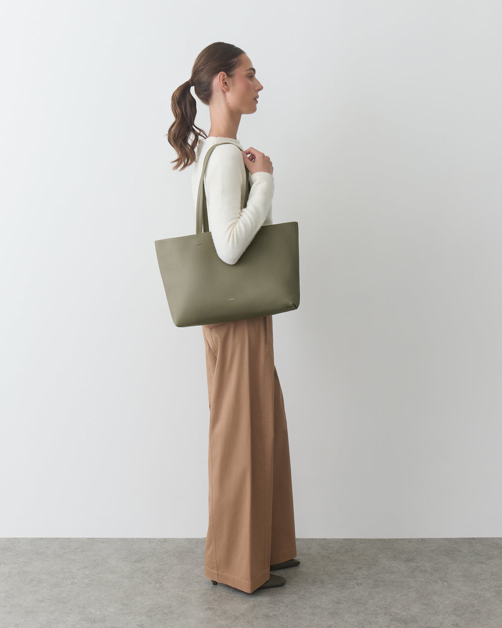Person standing with a tote bag, wearing long-sleeve top and wide-legged pants.