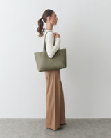 Person standing with a tote bag, wearing long-sleeve top and wide-legged pants.