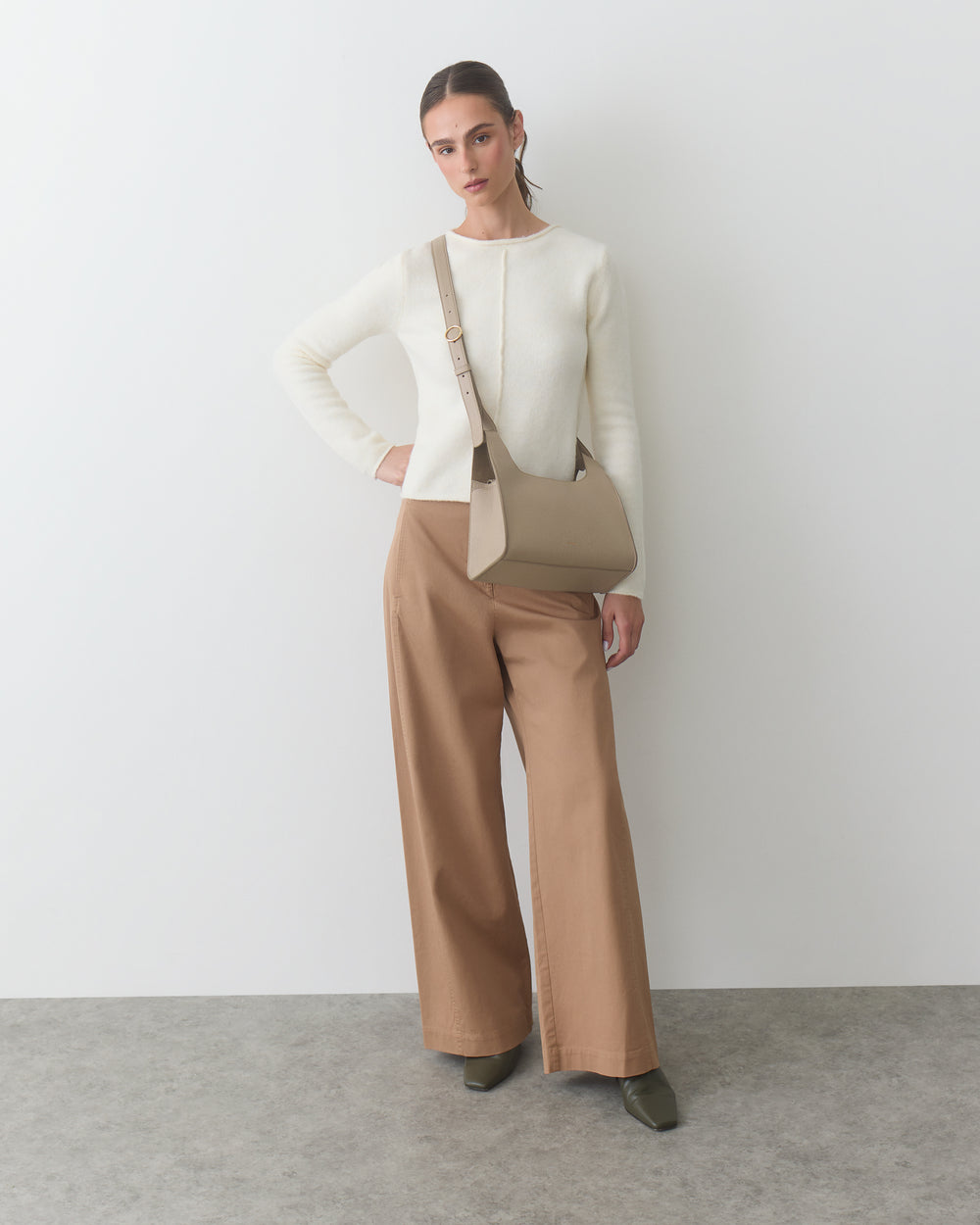 Person standing against wall holding a handbag, wearing a long-sleeve top and wide-leg pants.