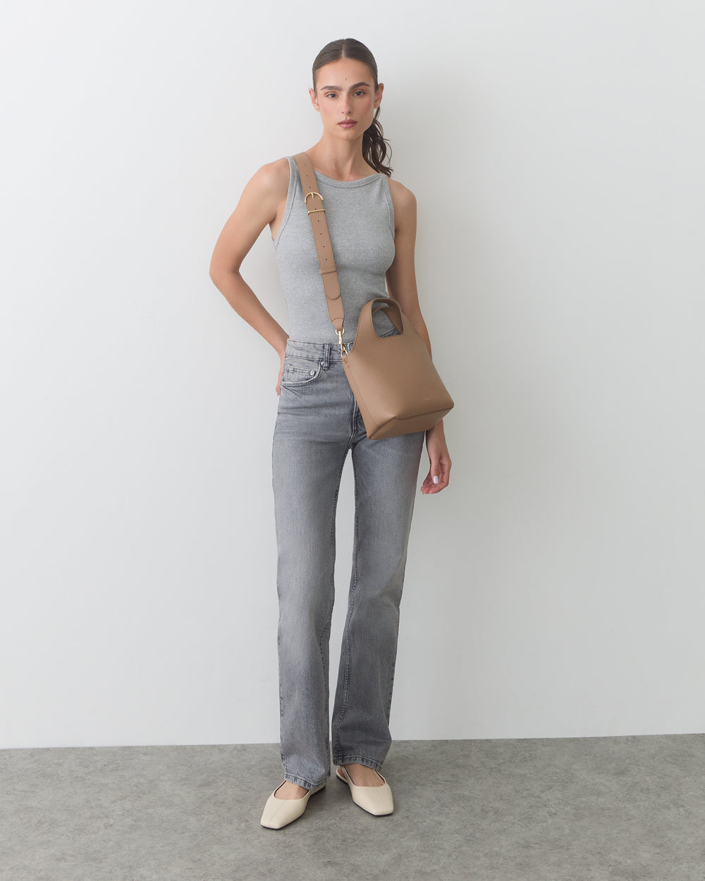 Person wearing tank top, jeans, holding small bag, standing against plain backdrop.