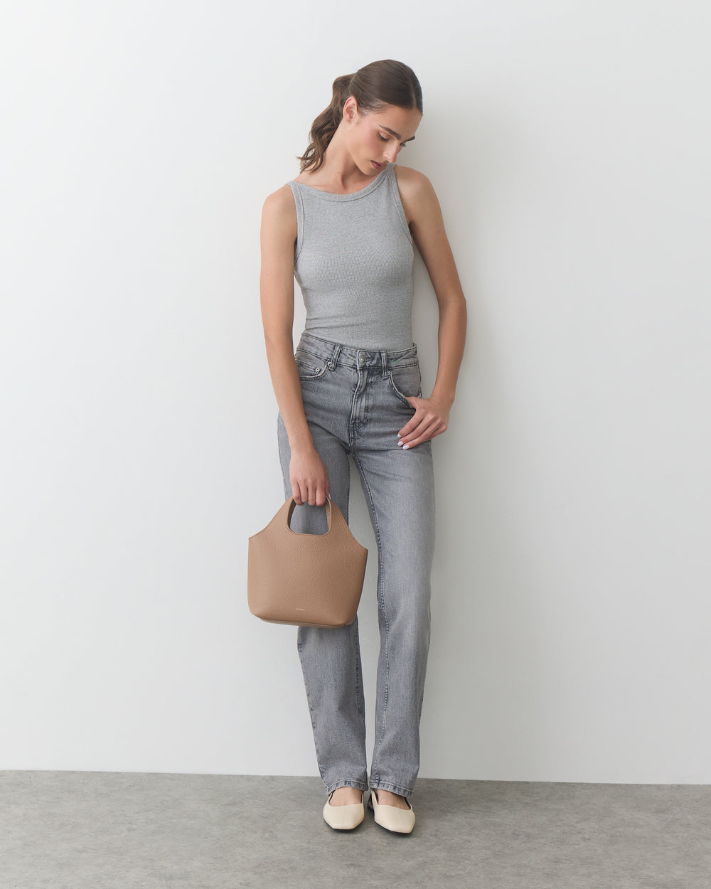 Person in tank top and jeans holding a handbag, standing against a plain background.