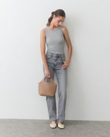 Person in tank top and jeans holding a handbag, standing against a plain background.
