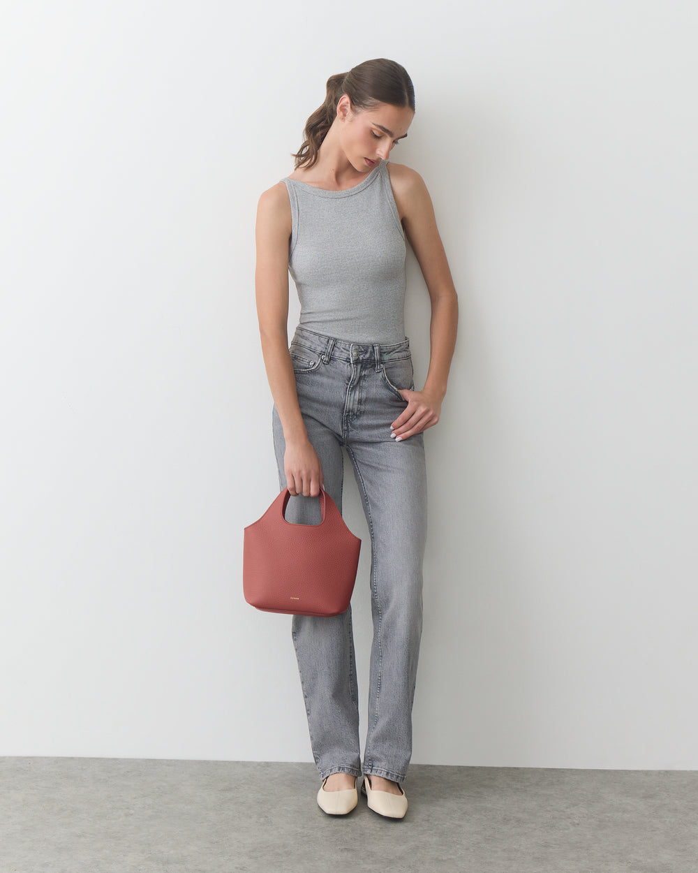 Person in tank top and jeans holding a handbag, standing against a plain backdrop.