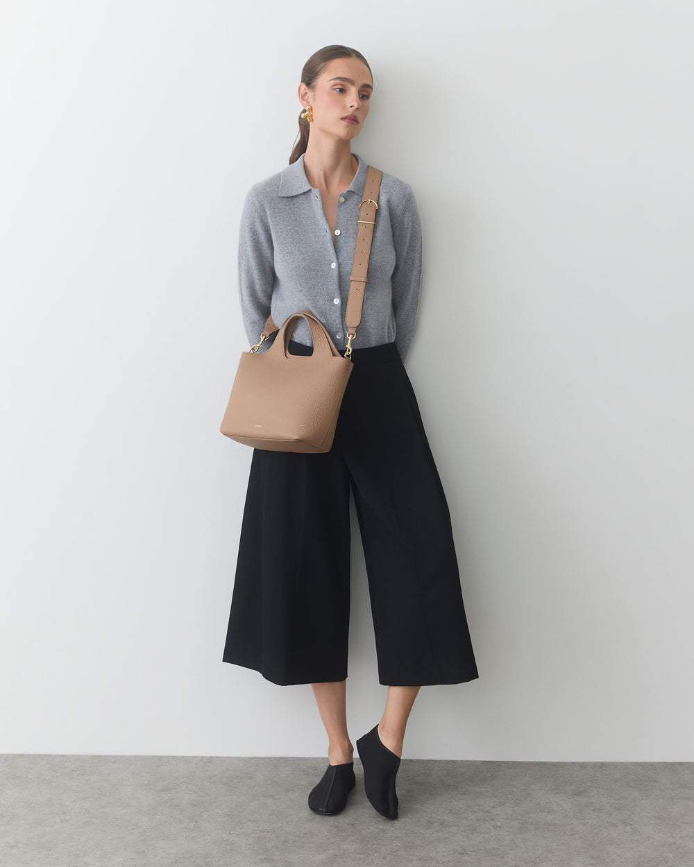Person leaning against a wall carrying a handbag with hands in pockets.