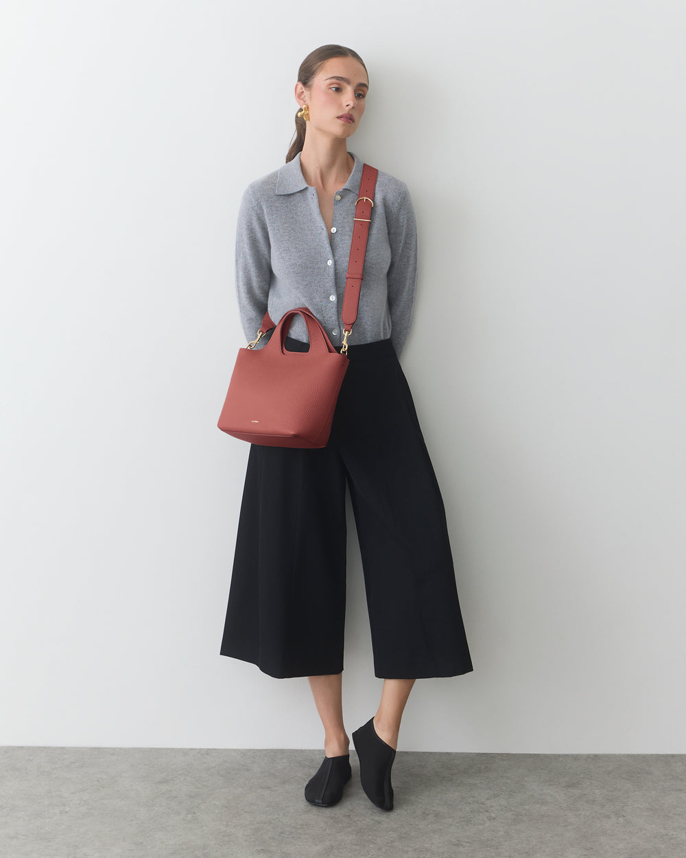 Person wearing a cardigan, culottes, holding a shoulder bag, leaning against a wall.