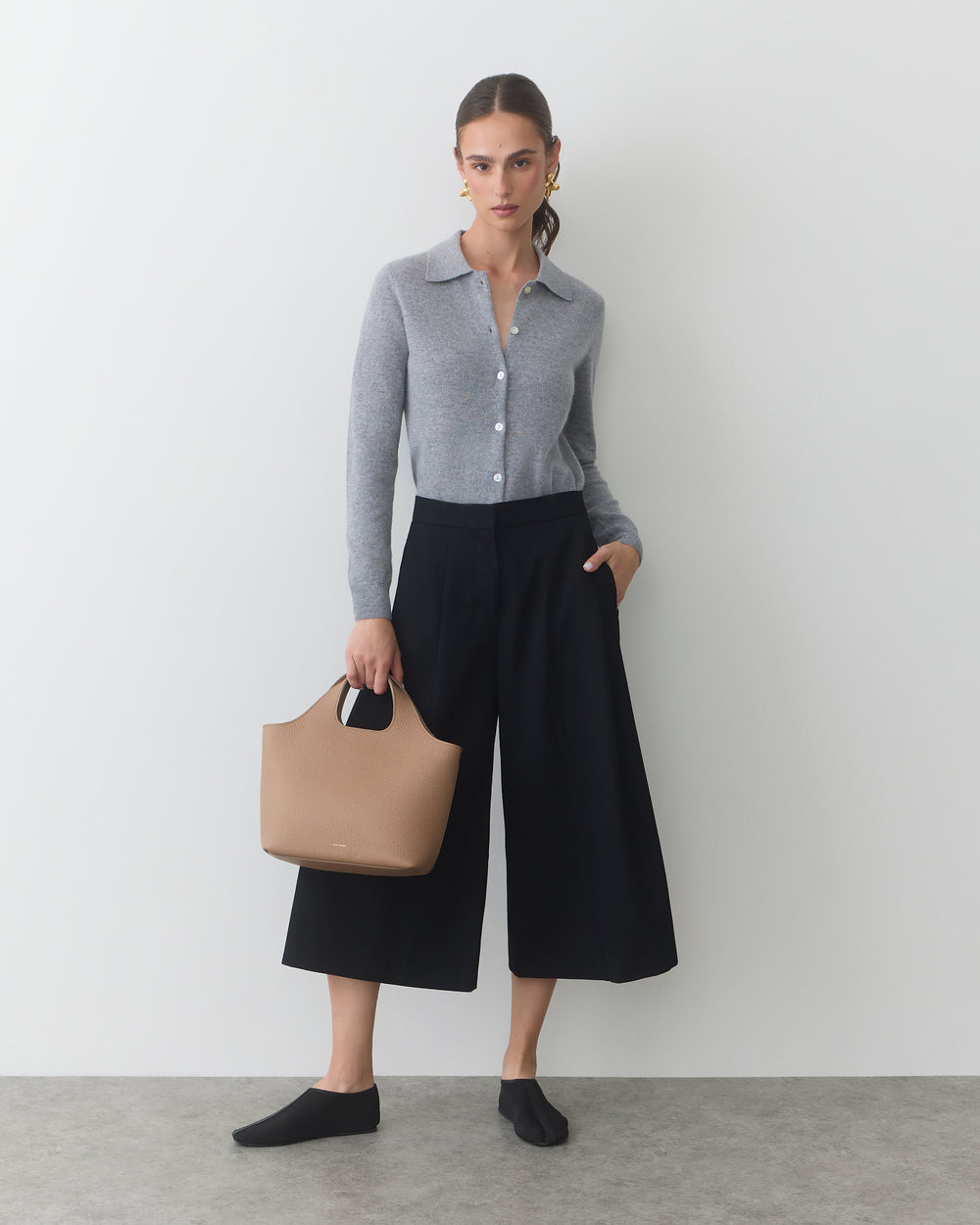 Person holding a handbag, wearing a long-sleeve top and wide-leg pants, standing indoors.