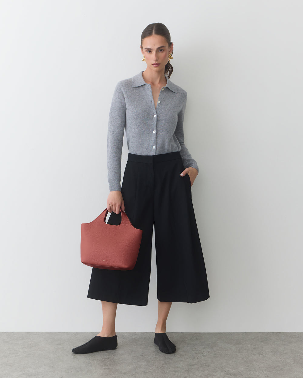 Person holding a handbag, wearing a long-sleeve top and wide-leg pants, standing indoors.