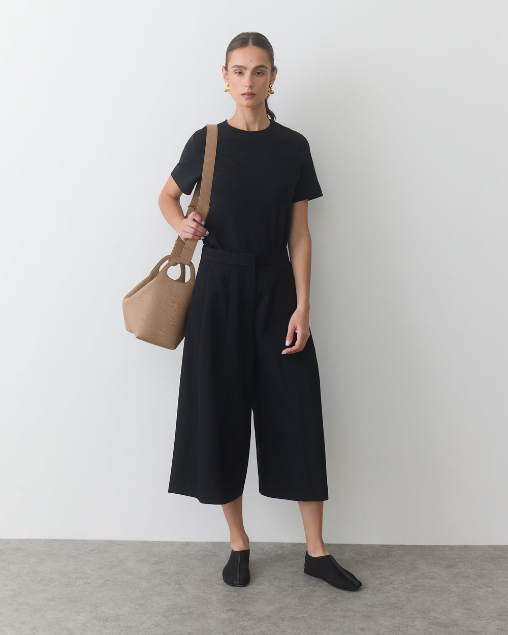 Person standing with a travel bag, wearing a short sleeve shirt and wide-leg pants.