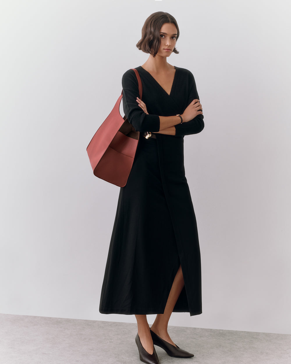 Person in long dress with shoulder bag and heels, standing against plain backdrop.