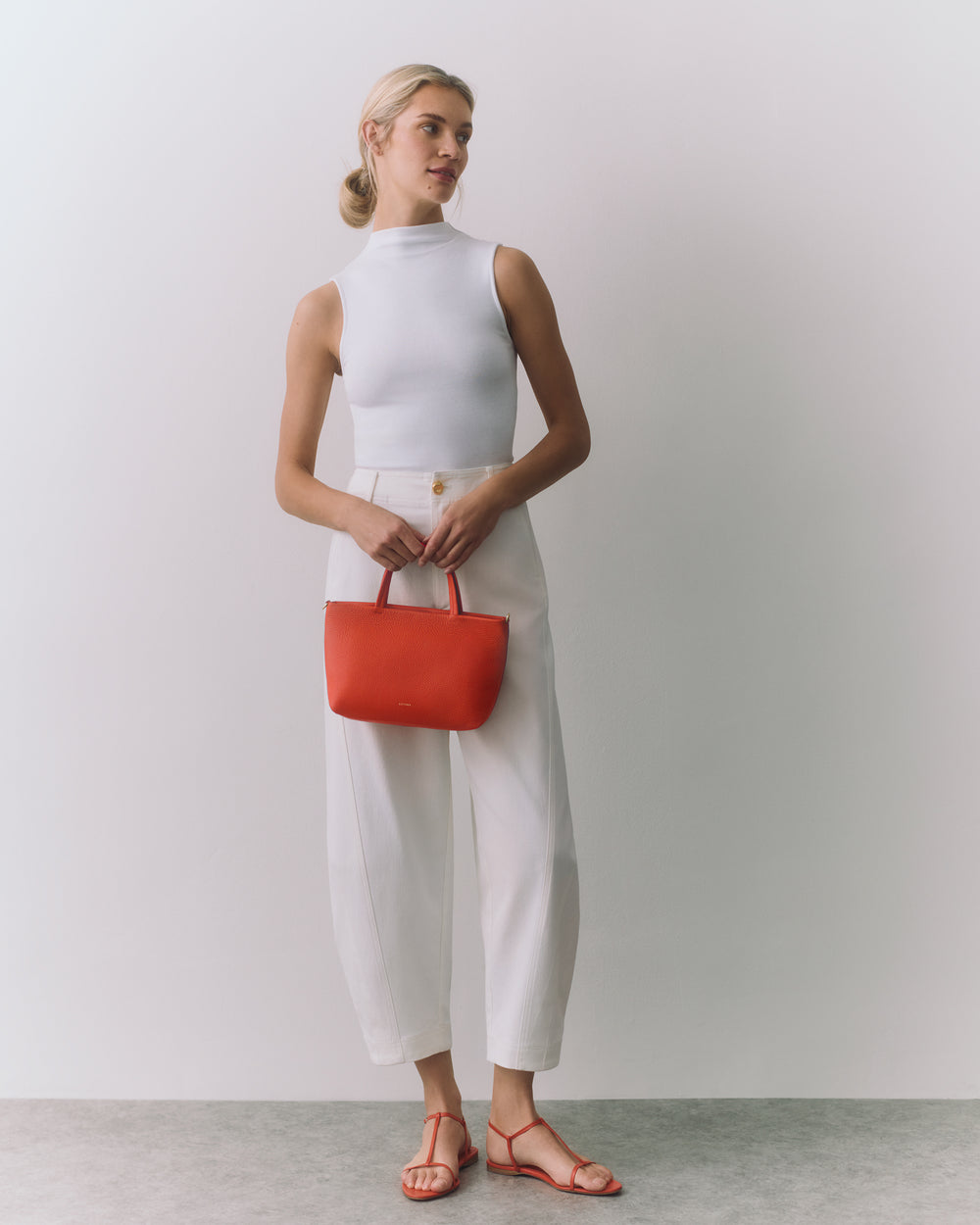 Woman in sleeveless top, pants, holding a handbag, wearing sandals.