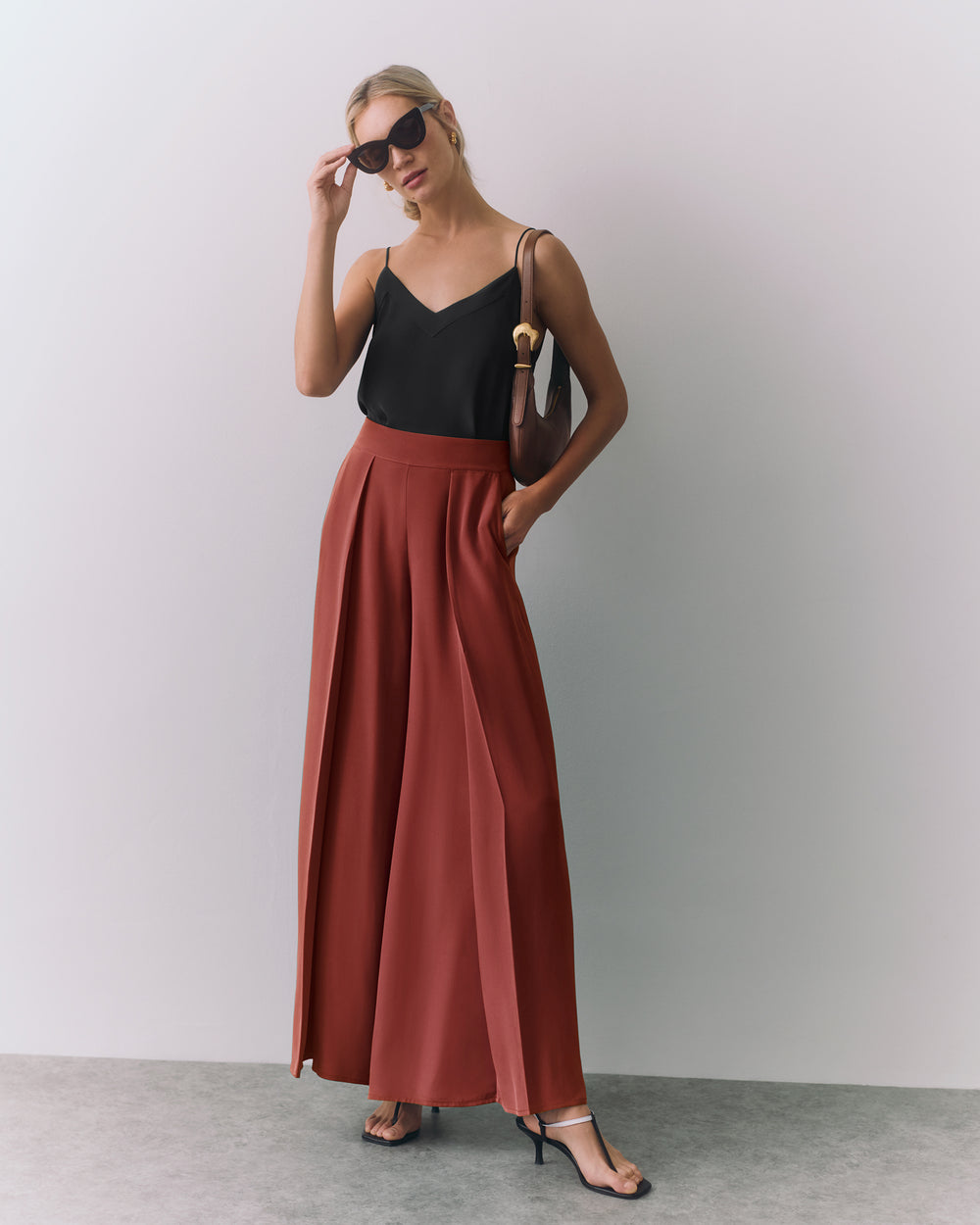 Person wearing sleeveless top and wide pants with sunglasses, holding a bag on shoulder.