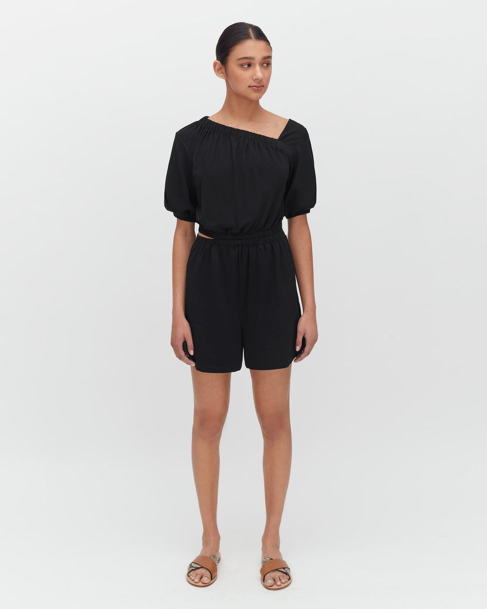 Person wearing a one-shoulder top and shorts with sandals, standing against a plain background.