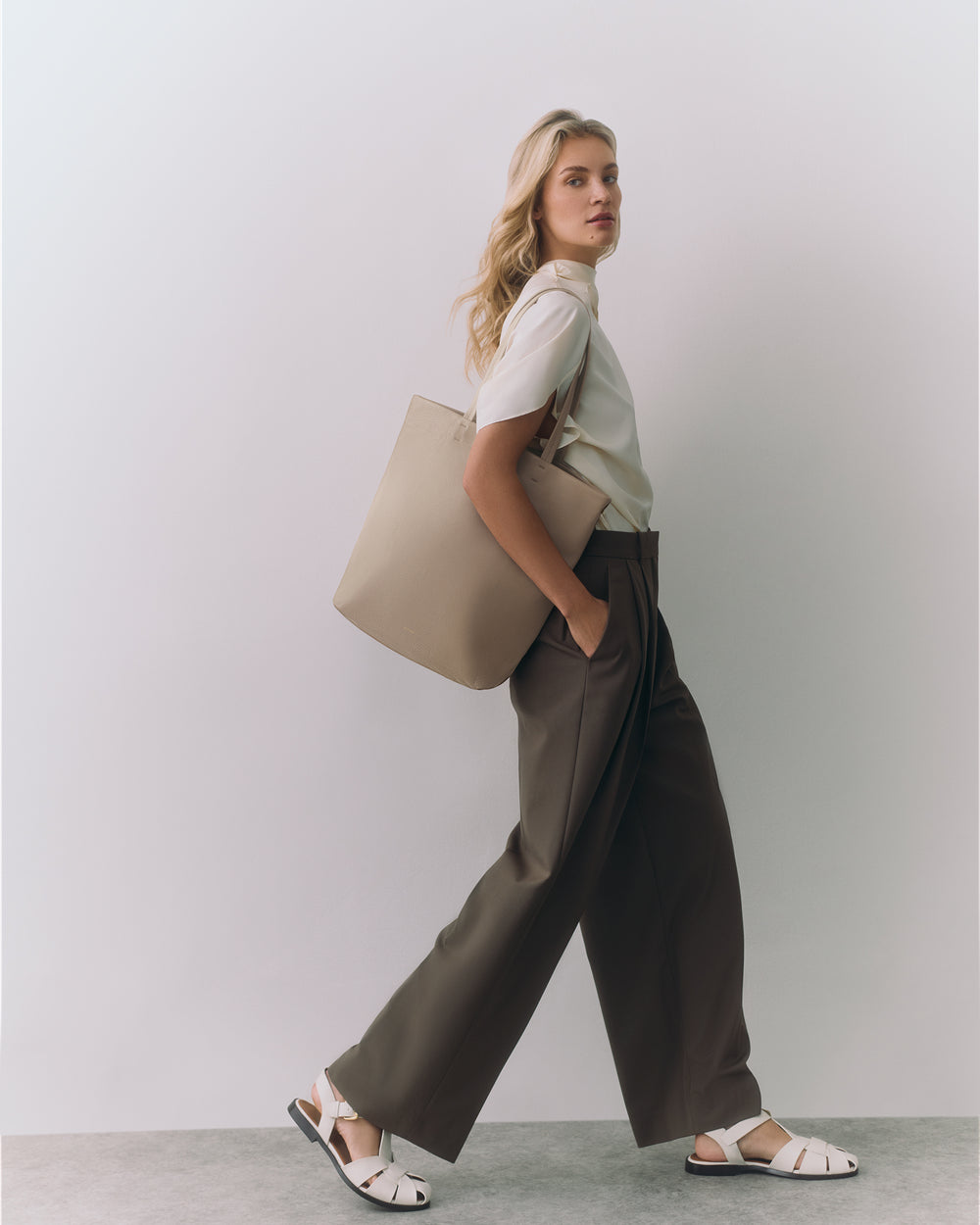 Person standing with hands in pockets, holding a bag, wearing sandals and wide-leg pants.