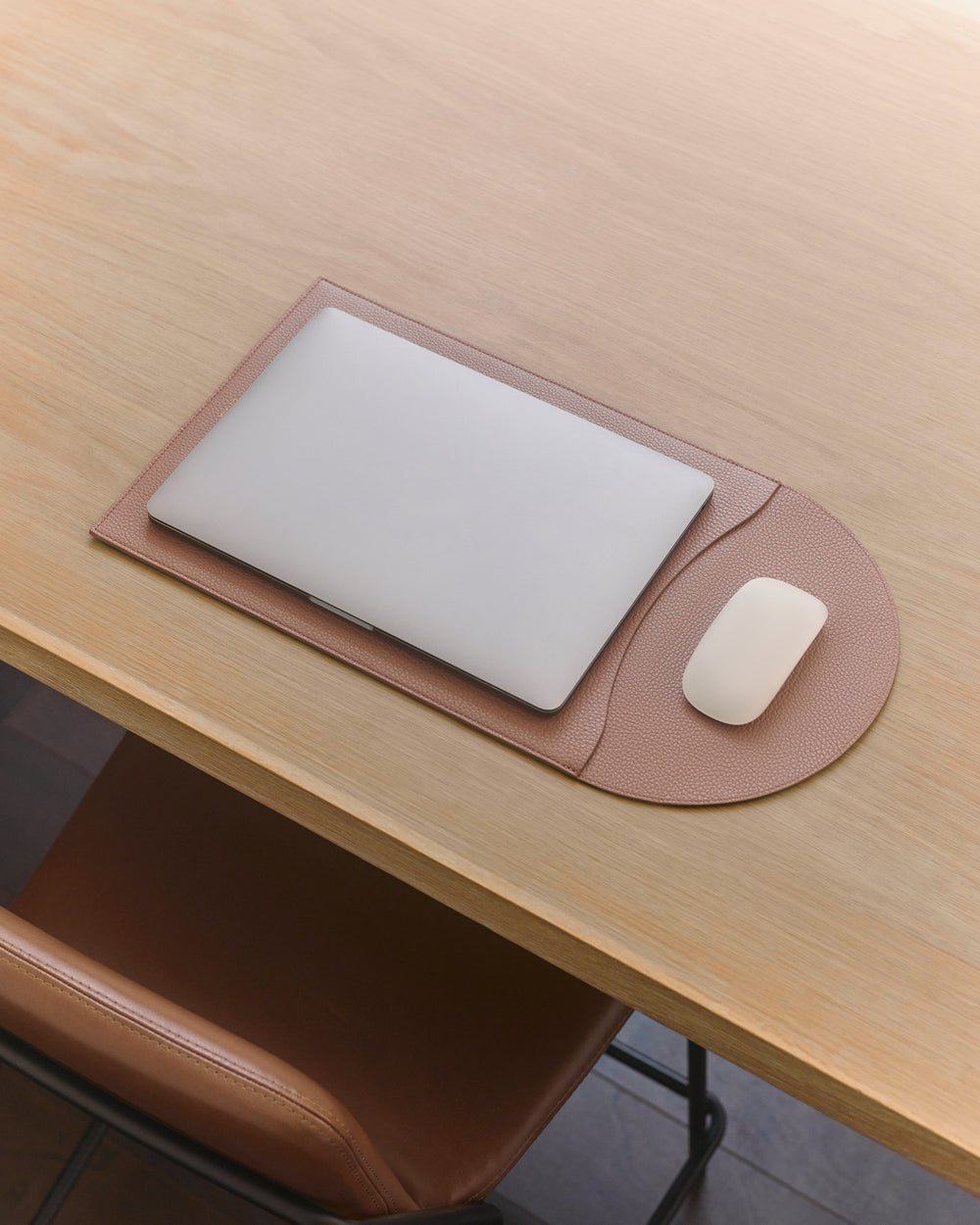 Laptop and mouse on a deskpad on a wooden table with a chair nearby.