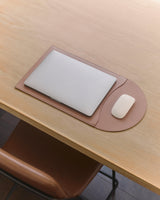 Laptop and mouse on a deskpad on a wooden table with a chair nearby.
