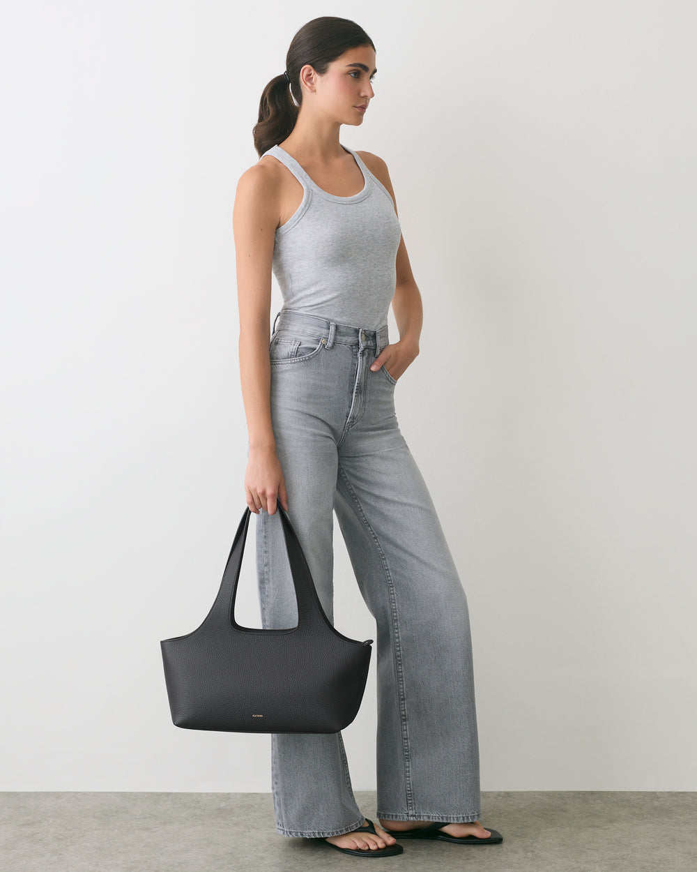 Person in tank top and jeans holding a handbag, standing against a white wall.