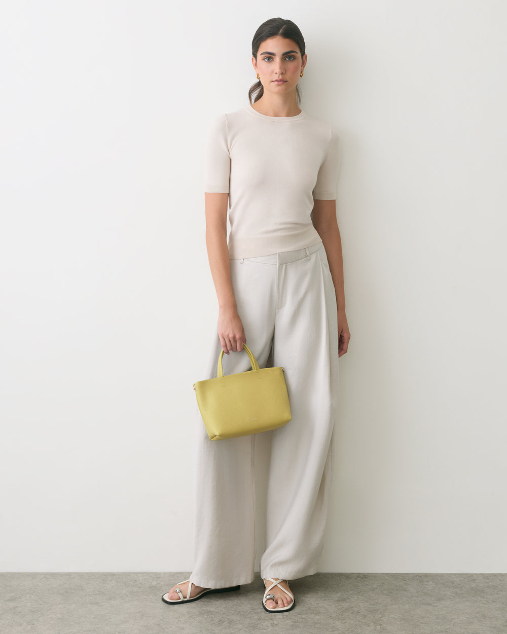 Person standing against wall holding a handbag wearing a short-sleeved top and wide-legged pants.