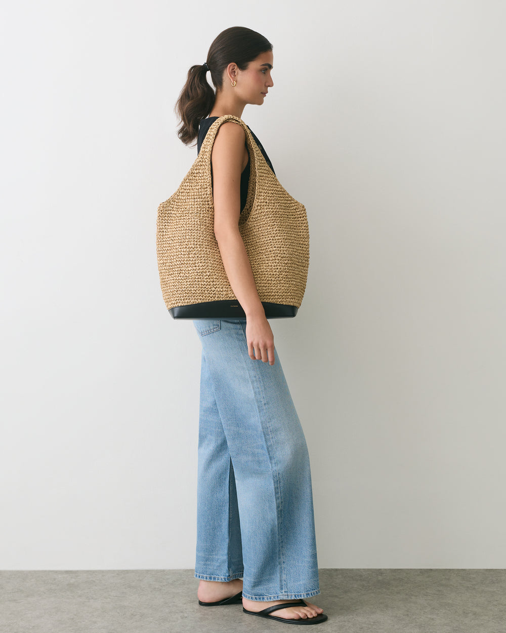 Person in wide-leg jeans, sleeveless top, with large woven bag and flip-flops, standing sideways.
