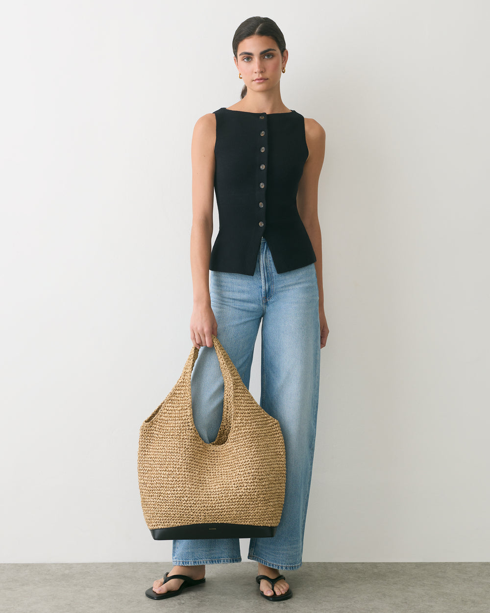 Person standing with tote bag, wearing sleeveless top, jeans, and sandals.
