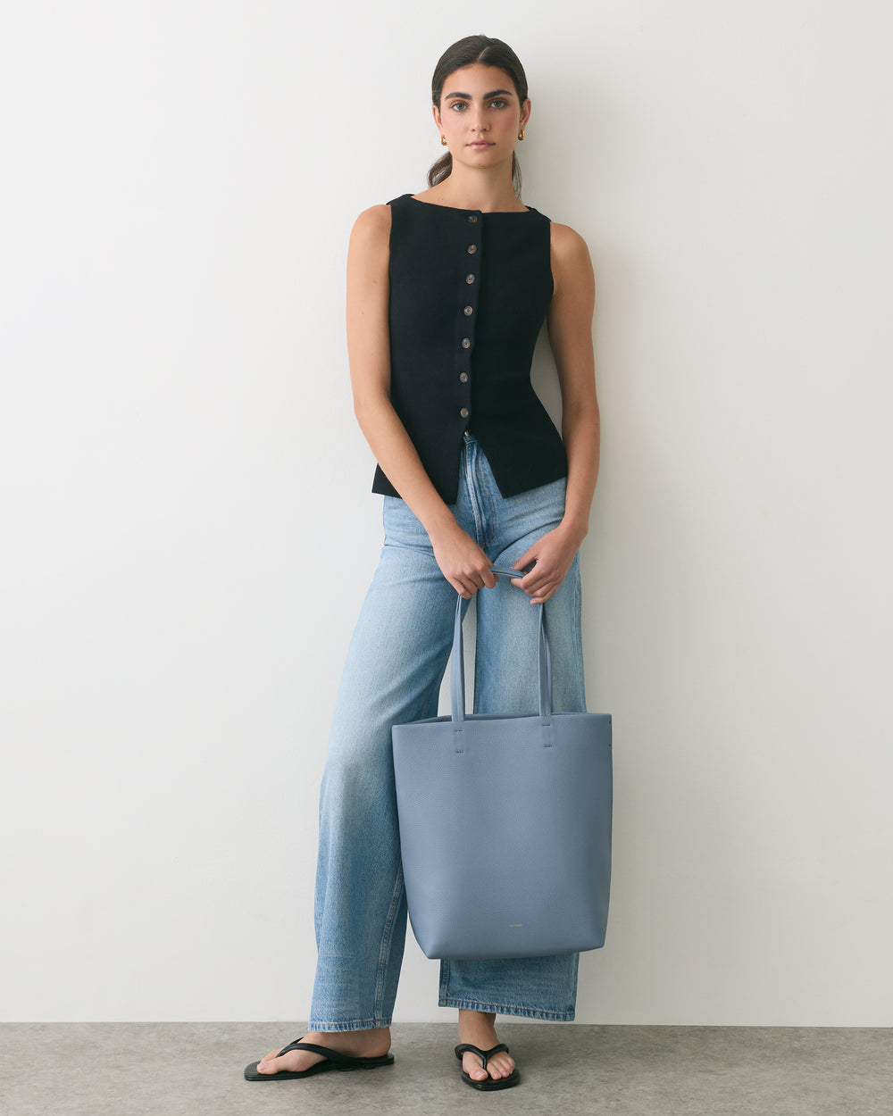 Person in sleeveless top and jeans holding a tote bag standing against a plain background.
