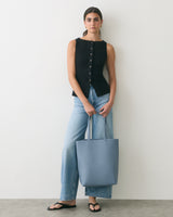 Person in sleeveless top and jeans holding a tote bag standing against a plain background.