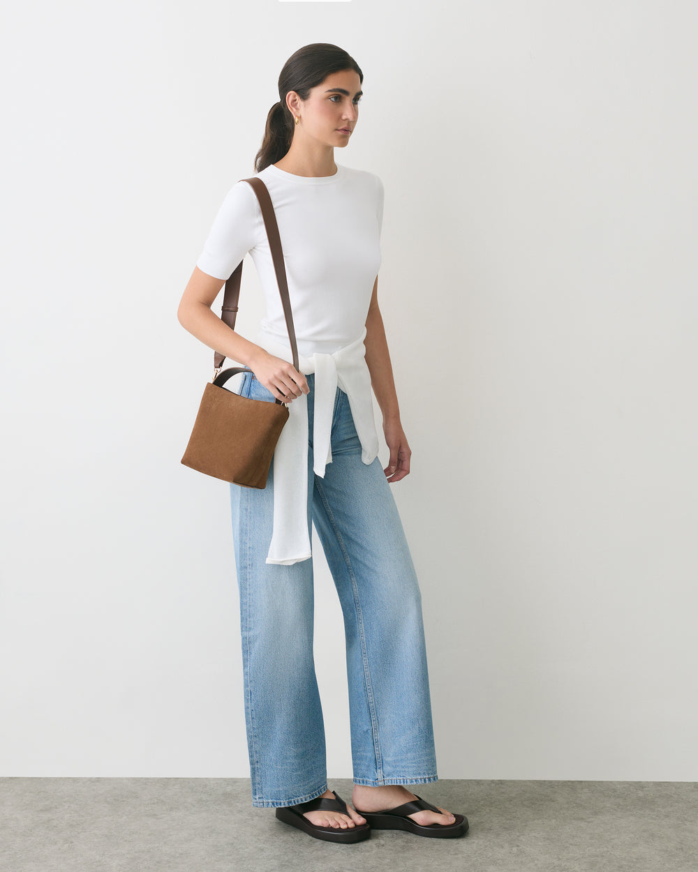 Person wearing a t-shirt, jeans, sandals, with a shoulder bag, standing against a plain wall.