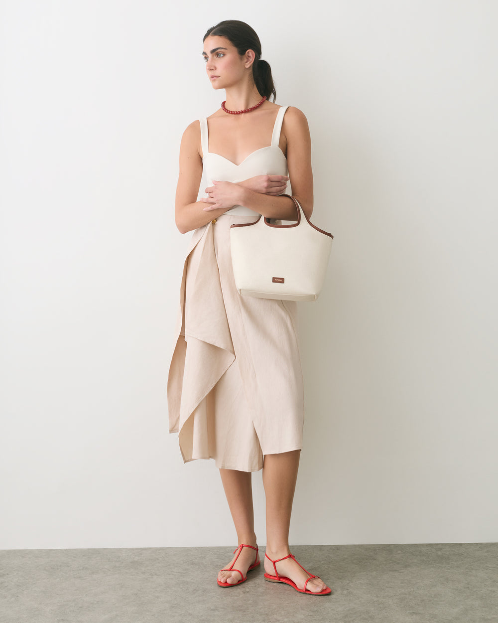 Person in a dress holding a handbag, wearing sandals and a necklace, standing against a plain wall.