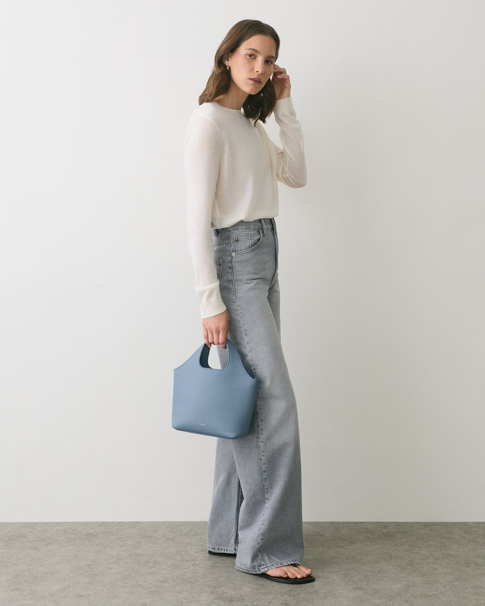 Person standing with jeans and a sweater holding a small handbag.
