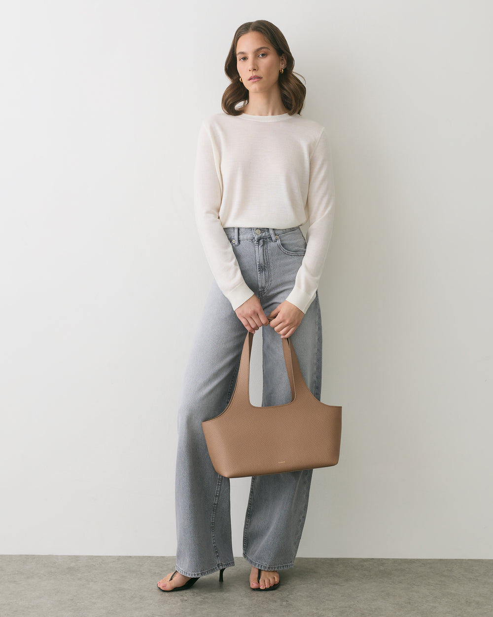 Person in sweater and jeans holding a handbag, standing against a plain backdrop.