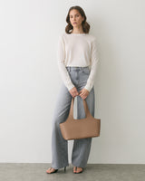 Person in sweater and jeans holding a handbag, standing against a plain backdrop.