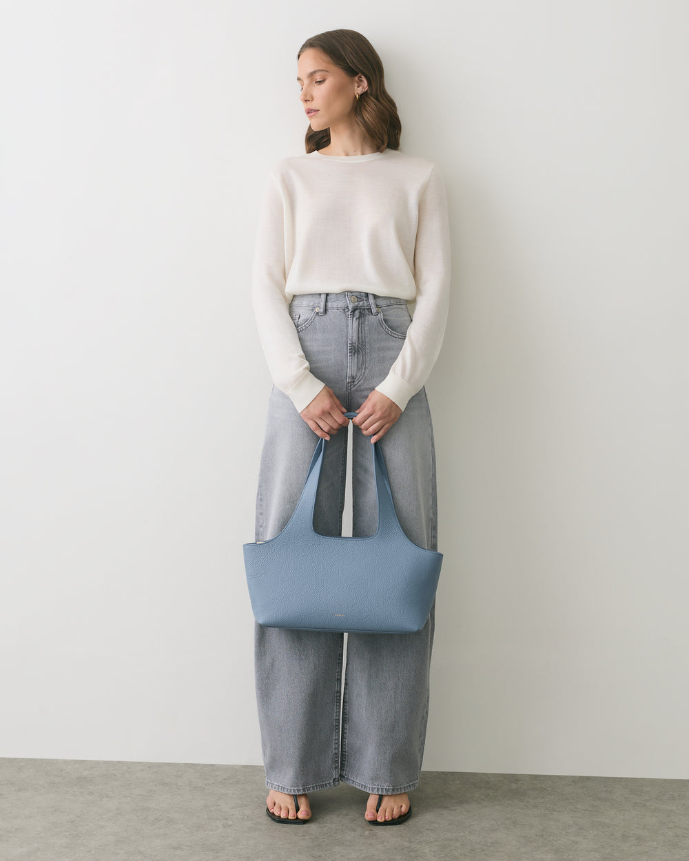 Person in sweater and jeans holding a handbag, standing against plain background.