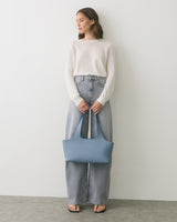 Person in sweater and jeans holding a handbag, standing against plain background.