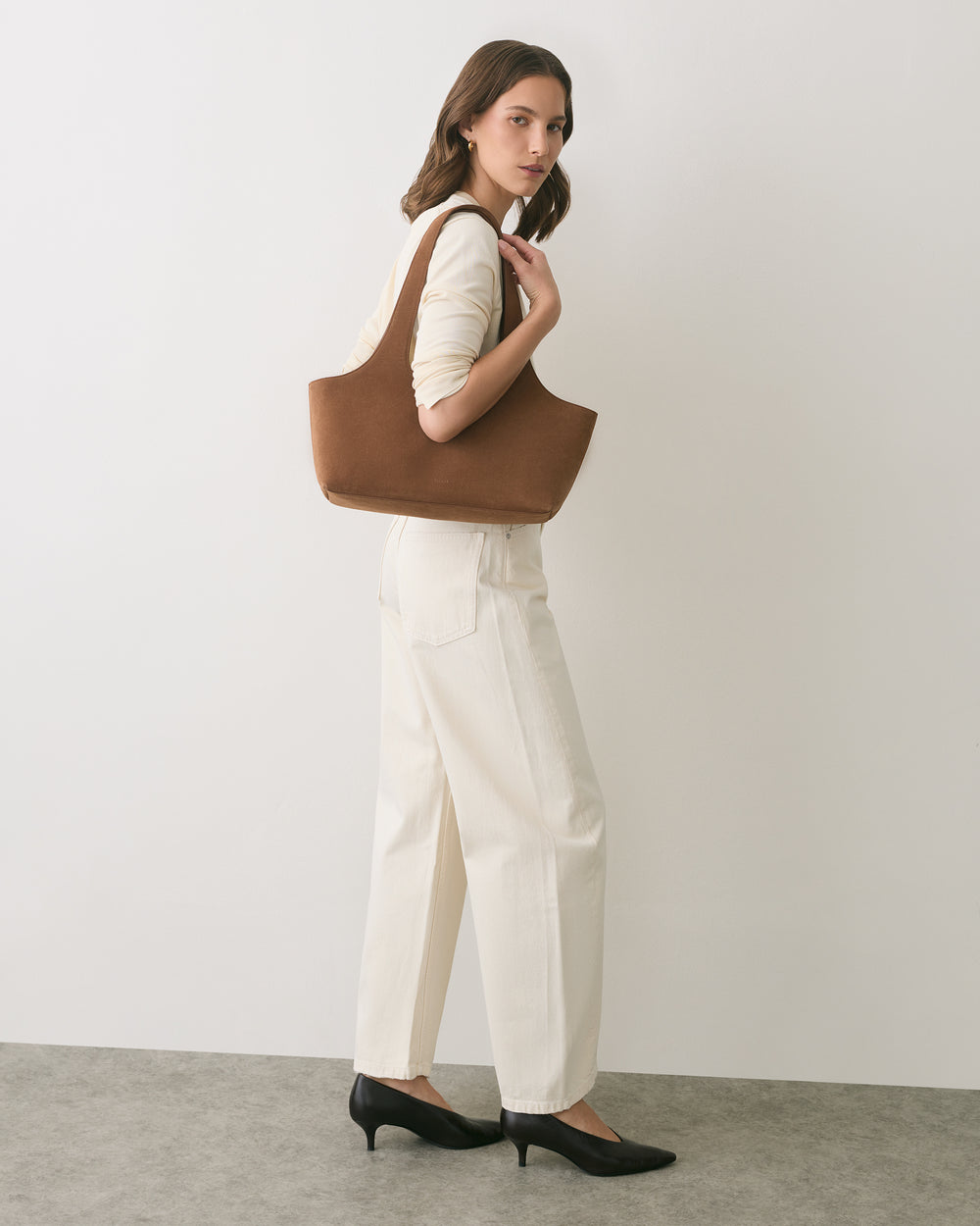 Person standing with shoulder bag, wearing long-sleeved top and pants with heels.