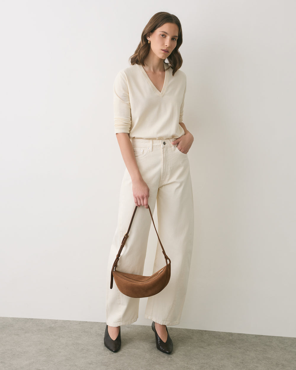 Person in a v-neck sweater and pants holding a shoulder bag, standing against a plain backdrop.
