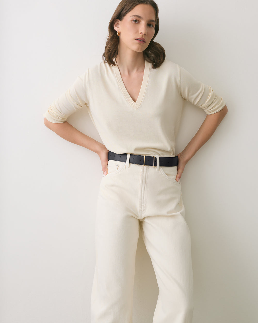 Person standing against a wall wearing a long-sleeve shirt and pants with hands on hips.