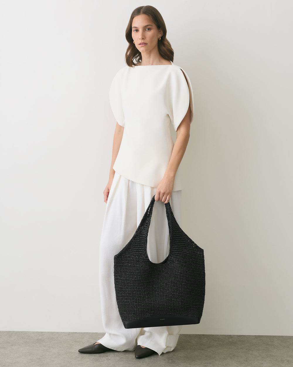 Person standing with a woven bag, wearing a sleeveless top and wide-leg pants.