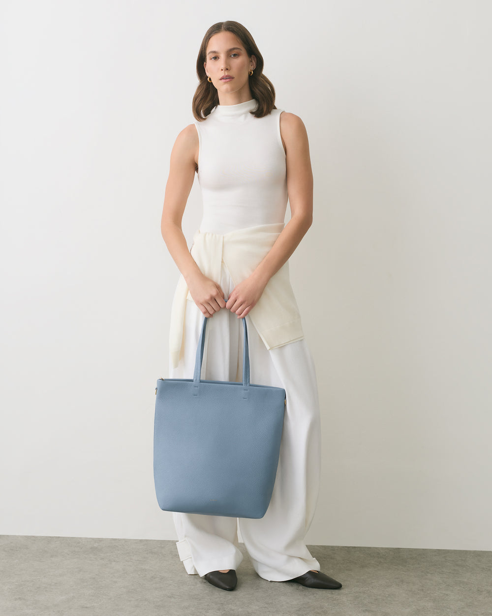 Person in sleeveless top holding a tote bag, wearing wide-leg pants.