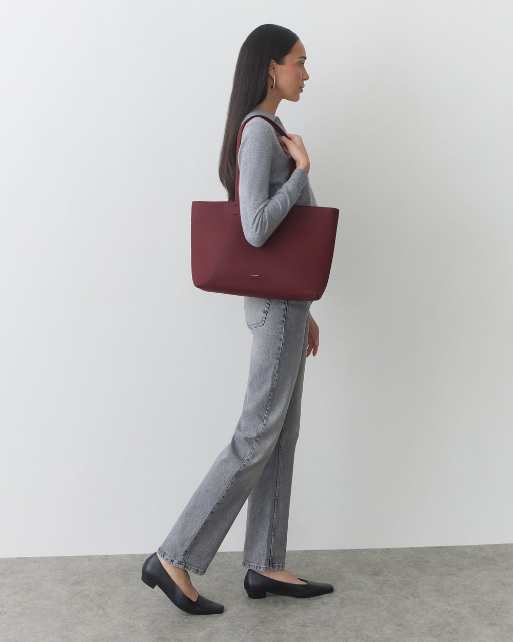 Person holding large tote bag, wearing sweater and jeans, standing sideways.