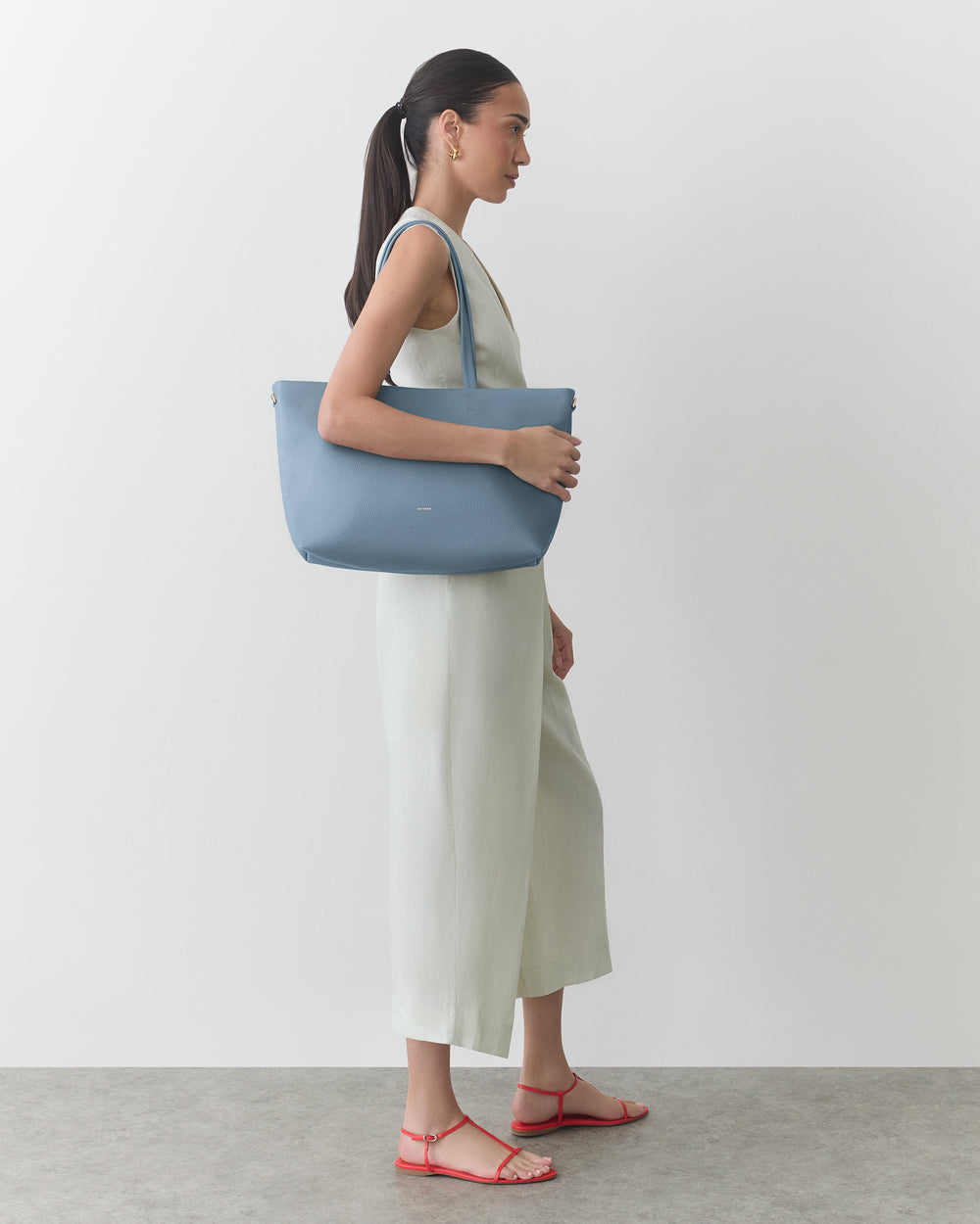 Person standing, holding a shoulder bag, wearing a sleeveless dress and sandals.