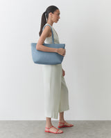 Person standing, holding a shoulder bag, wearing a sleeveless dress and sandals.