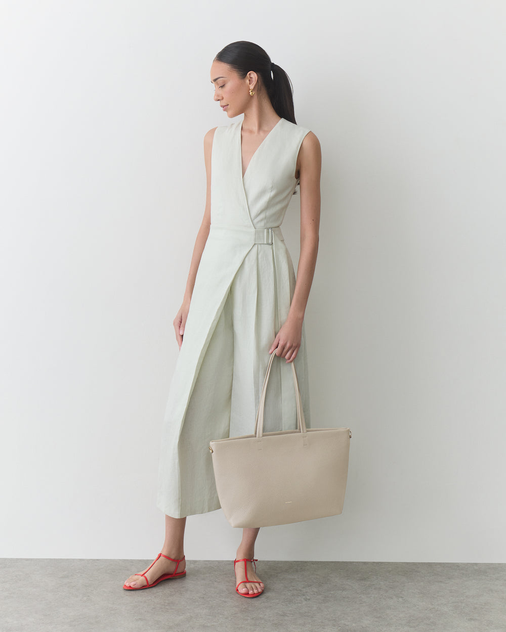 Person in sleeveless dress holding a large handbag, wearing sandals, standing on a plain background.