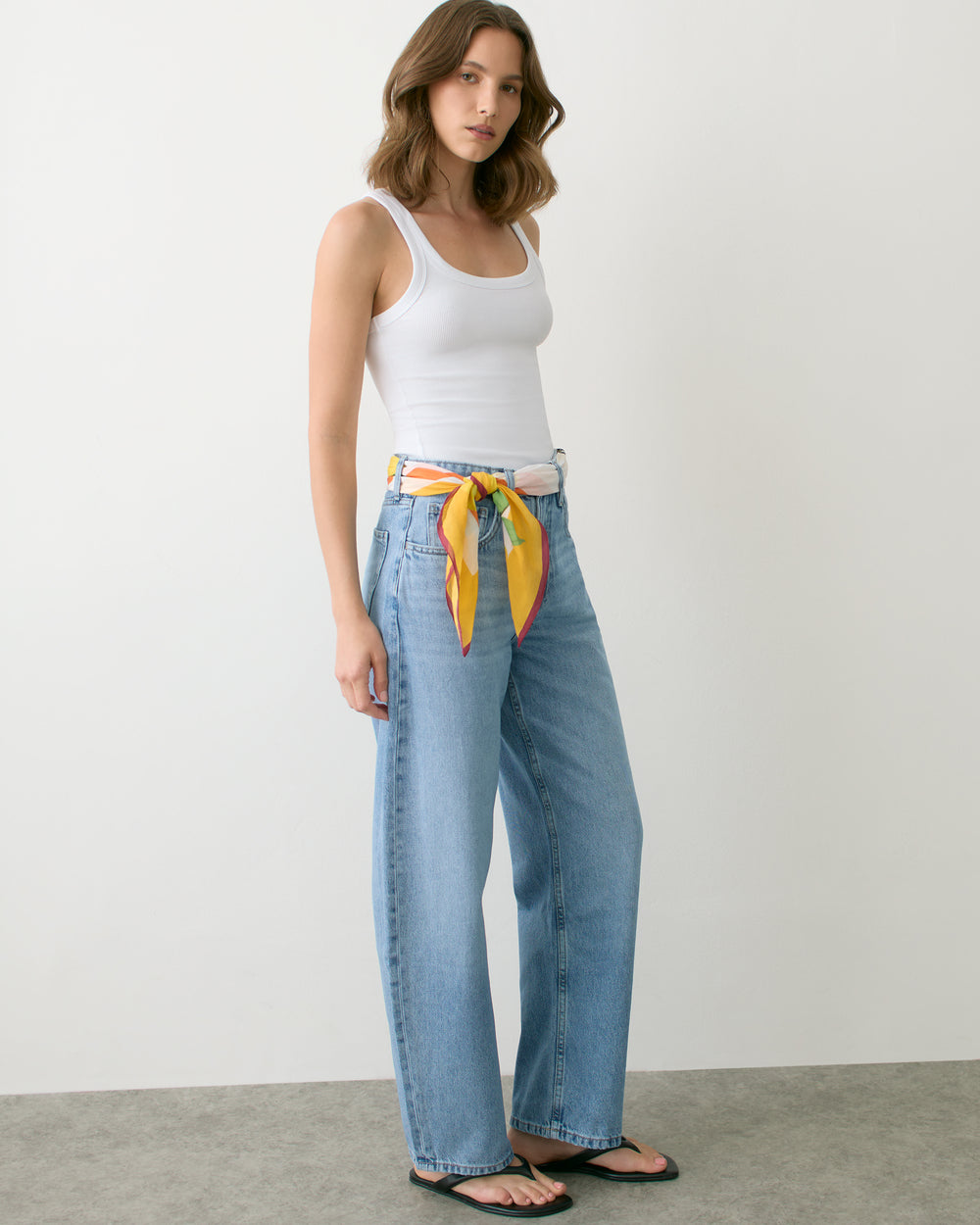 Person in tank top and jeans with a scarf as a belt standing against a plain backdrop.