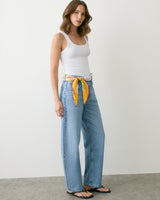 Person in tank top and jeans with a scarf as a belt standing against a plain backdrop.