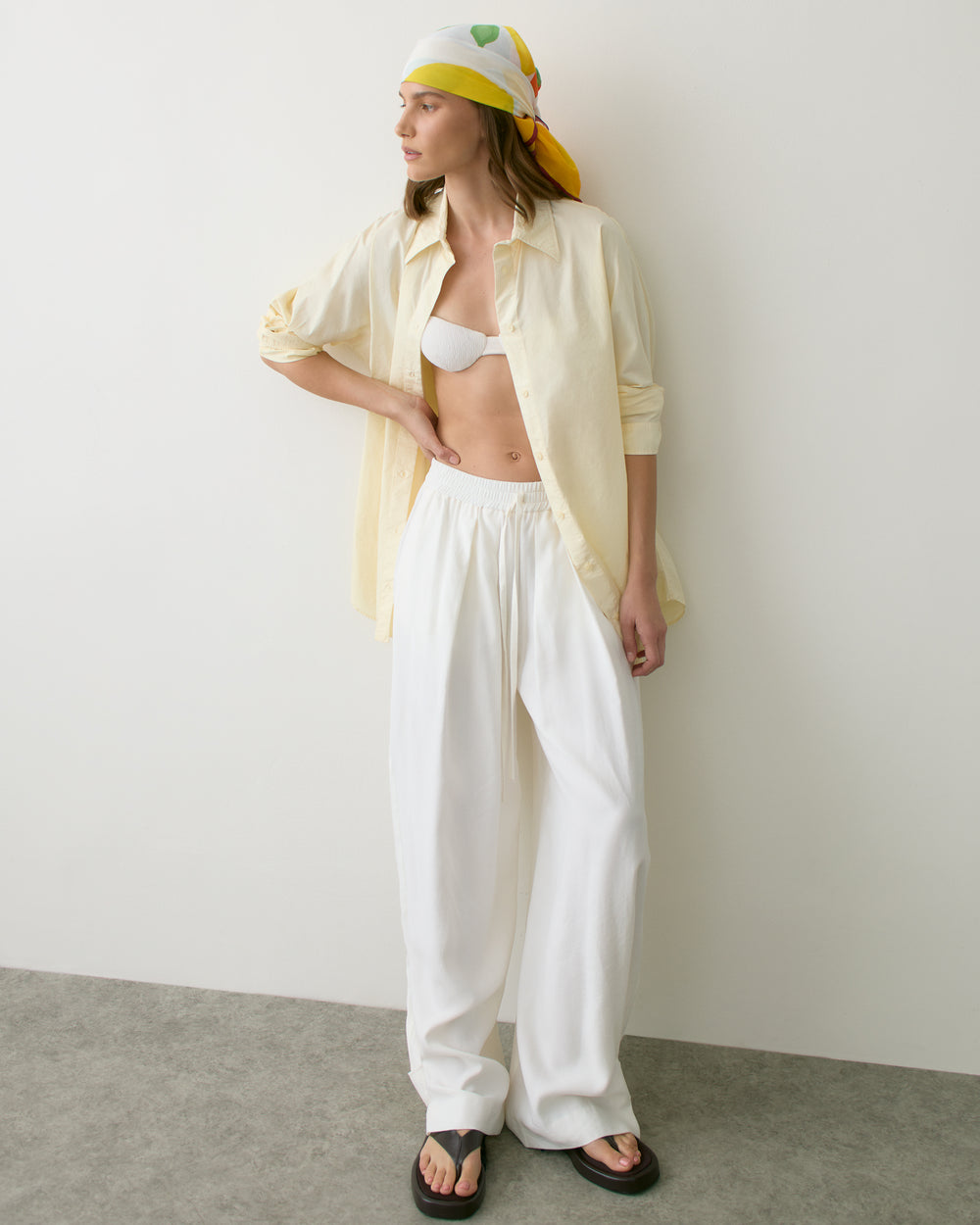 Person wearing a headscarf, open shirt, bikini top, wide pants, and sandals, standing against a wall.
